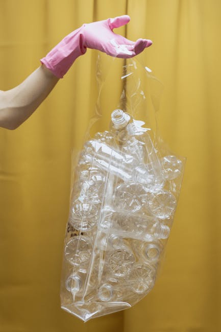 A person wearing a white t-shirt and disposable gloves is holding a white plastic bin labeled 'PLASTIC' in black marker. The bin contains a black plastic rubbish bag that is open and draped over the s