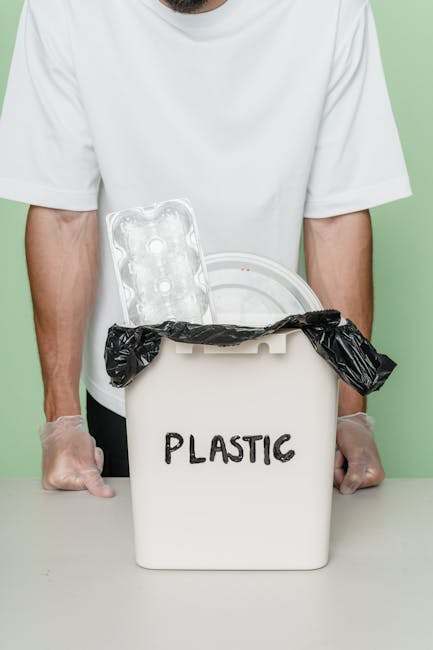 A close-up view of multiple black plastic rubbish bags filled with waste, stacked against a plain, light grey wall. The bags are made of shiny, smooth plastic with visible creases and folds, indicatin