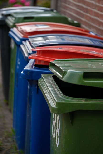 A row of large outdoor wheelie bins with varying colours, including green, red, and blue, positioned along a pavement adjacent to a brick wall. The green bin in the foreground has a textured lid with parallel ridges, while the blue and red bins have smooth, slightly reflective surfaces on their lids. The bins are tightly lined up, with their front faces visible, showing their sturdy plastic construction. The background includes a blurred view of another bin and hints of greenery, suggesting these are placed outside residential or commercial premises for waste collection. The environment appears clean and well-maintained, indicative of a typical outdoor rubbish storage area for waste disposal or alternative collection services like those offered by Rubbish Collection London, which specializes in private waste handling and on-site clearance solutions, complementing traditional rubbish collection methods.