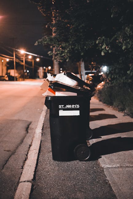 A black wheeled rubbish bin marked with the text 'ST. JOHN'S' positioned on a paved sidewalk beside a street at night. The bin is filled with various types of waste, including cardboard boxes, paper, and plastic, some of which are protruding and stacked unevenly. The background features dim street lighting, illuminated by nearby street lamps, with trees and parked cars visible along the roadside. The environment appears quiet and residential, with the bin placed close to the curb for potential collection. The scene suggests an area where residents or private waste services, such as Rubbish Collection London, may handle refuse outside traditional municipal collection points, supporting the concept of alternative waste disposal methods and on-site rubbish management.
