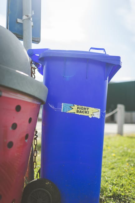 A row of large outdoor wheelie bins with varying colours, including green, red, and blue, positioned along a pavement adjacent to a brick wall. The green bin in the foreground has a textured lid with parallel ridges, while the blue and red bins have smooth, slightly reflective surfaces on their lids. The bins are tightly lined up, with their front faces visible, showing their sturdy plastic construction. The background includes a blurred view of another bin and hints of greenery, suggesting these are placed outside residential or commercial premises for waste collection. The environment appears clean and well-maintained, indicative of a typical outdoor rubbish storage area for waste disposal or alternative collection services like those offered by Rubbish Collection London, which specializes in private waste handling and on-site clearance solutions, complementing traditional rubbish collection methods.